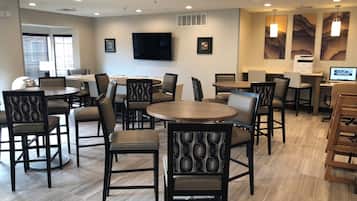 A hotel dining area with round tables and chairs, a flat-screen TV mounted on the wall, and artwork on the walls.