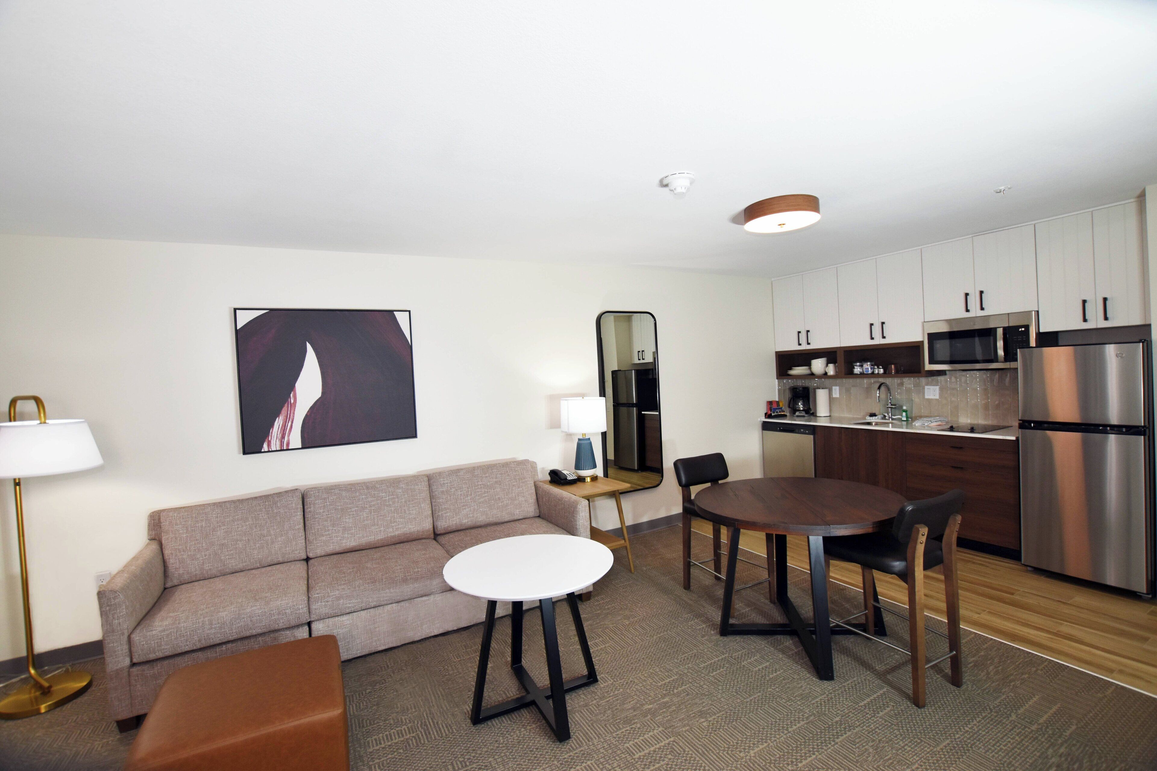 A modern hotel room with a sofa, a round dining table, a small kitchen area with a microwave and refrigerator, and a large mirror.