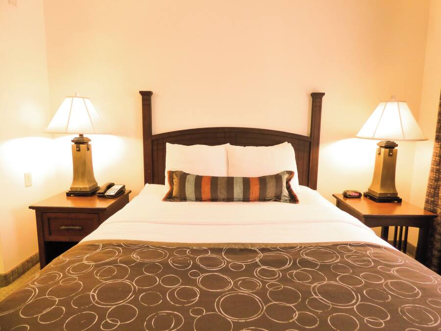 A hotel room with a bed, two bedside tables with lamps, a nightstand, and a clock.
