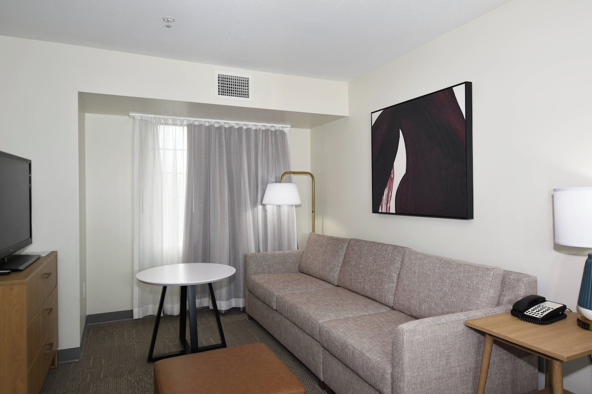 Ett hotellrum med en soffa, ett litet runt bord, en tv, en lampa och en tavla på väggen.