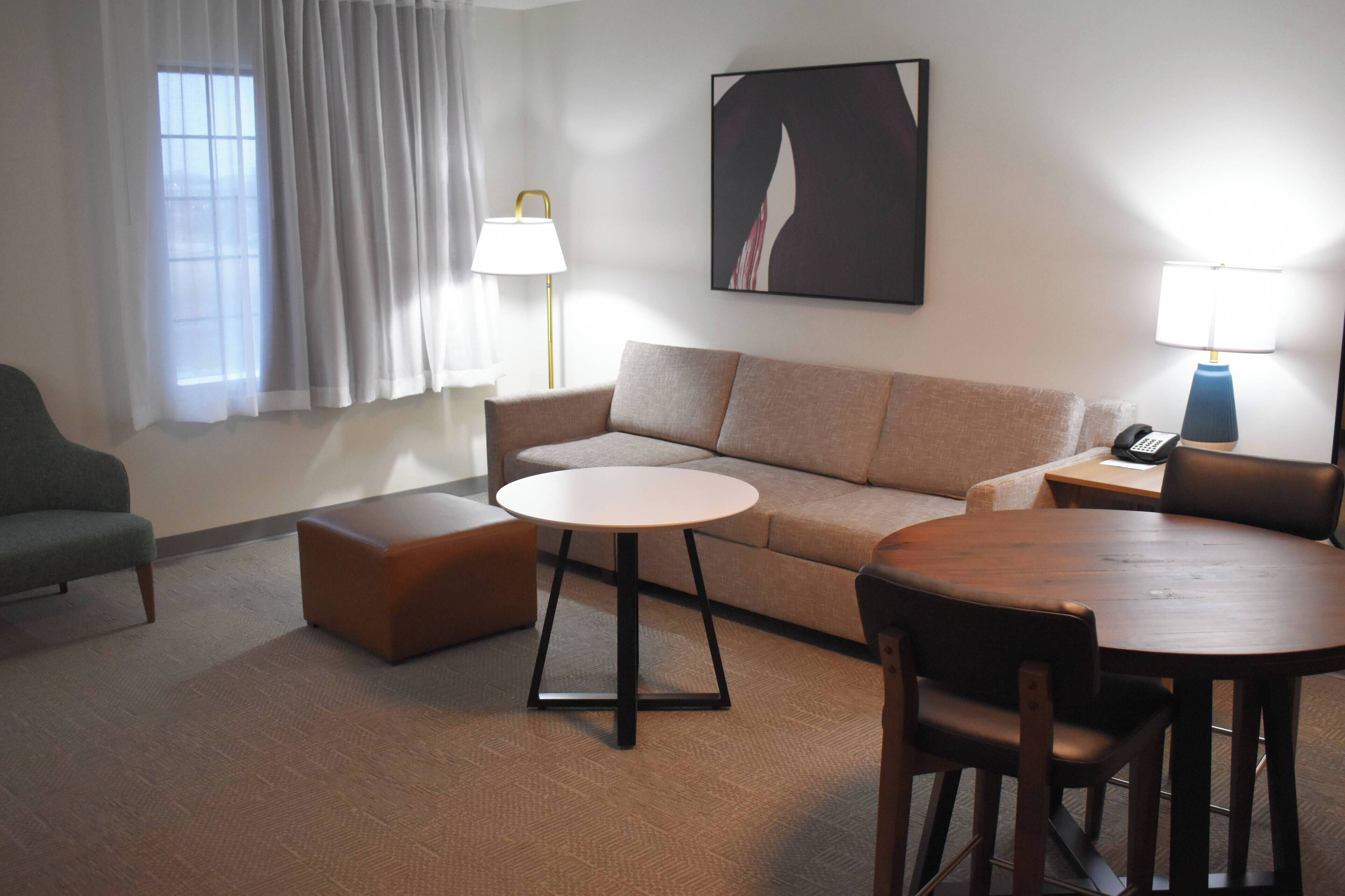 A hotel room with a sofa, a round coffee table, a dining table with chairs, a lamp, and a painting on the wall.