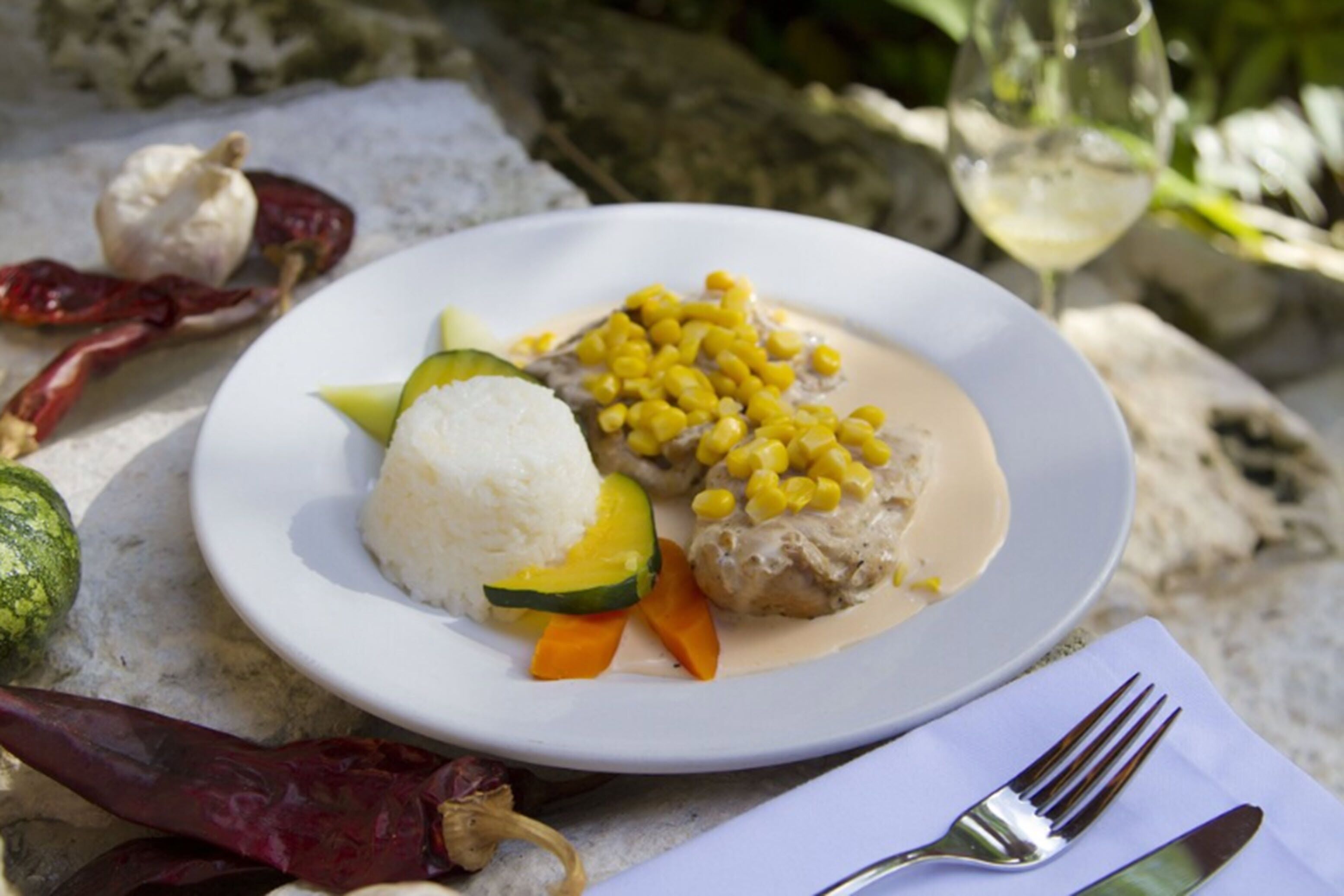 A plate of food with rice, vegetables, and a meat dish, accompanied by a glass of white wine and a fork on a napkin.