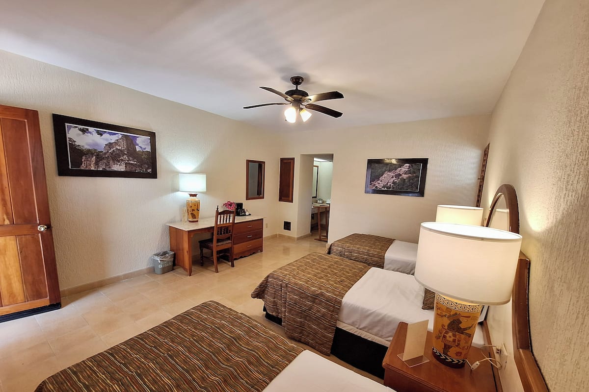 A hotel room with two beds, a desk, a chair, a ceiling fan, and two framed pictures on the walls.