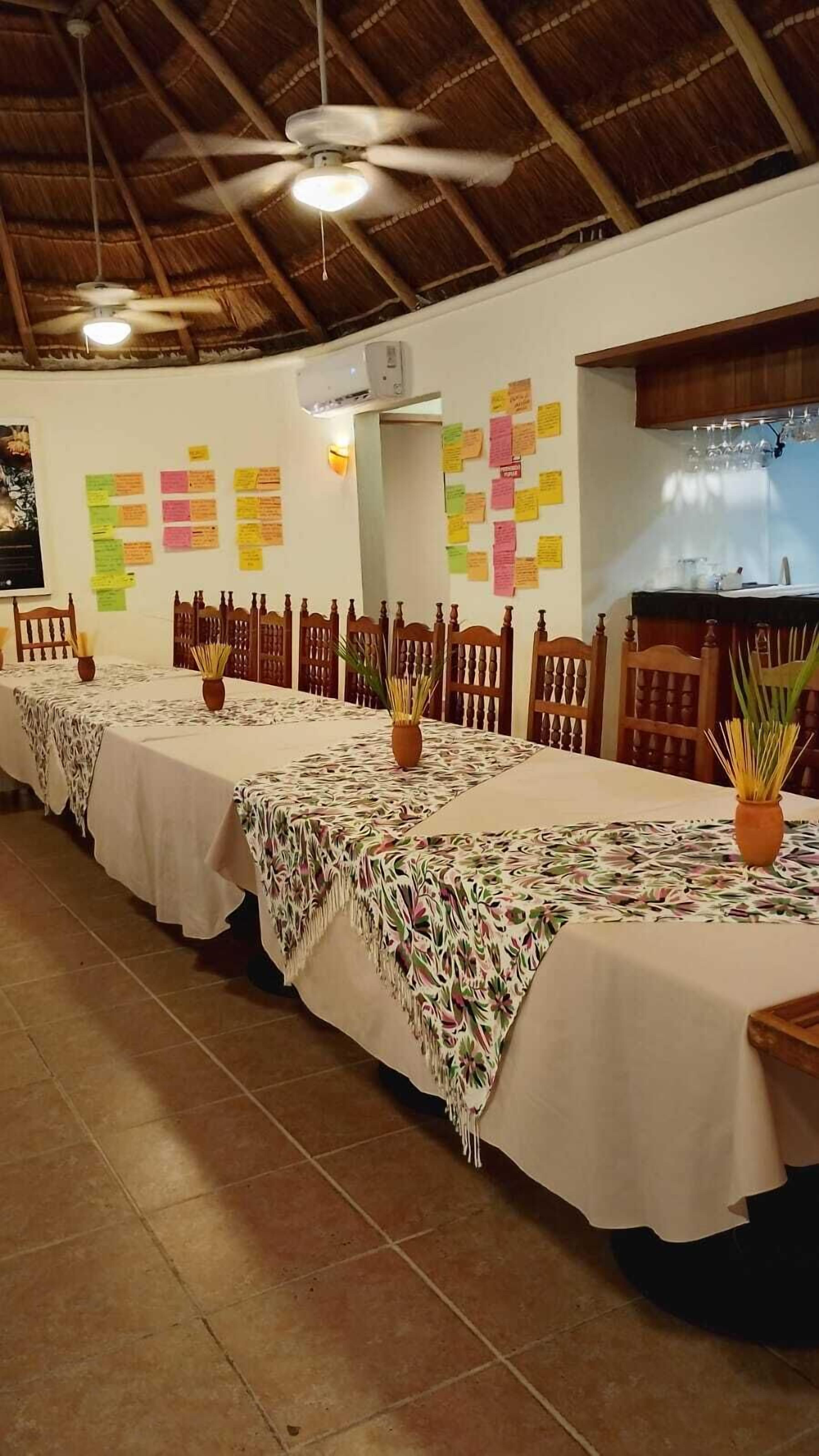 A long dining table with white tablecloths and potted plants, wooden chairs, and a ceiling fan in a room with a tiled floor and a wooden ceiling.