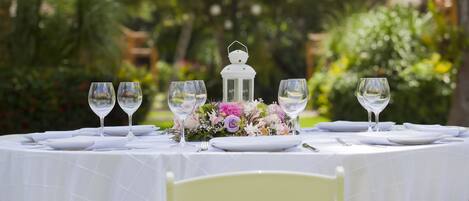 Mesa de comedor exterior con manteles blancos, platos y copas de vino, centrada con un farol y un arreglo floral.