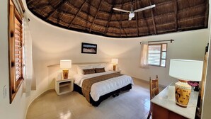 Un dormitorio con cama, dos mesitas de noche con lámparas, ventilador de techo, ventana con persianas y un cuadro en la pared.