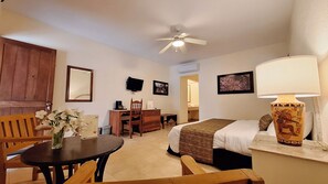 A hotel room with a bed, a desk, a chair, a table with a vase of flowers, a ceiling fan, and wall-mounted artwork.