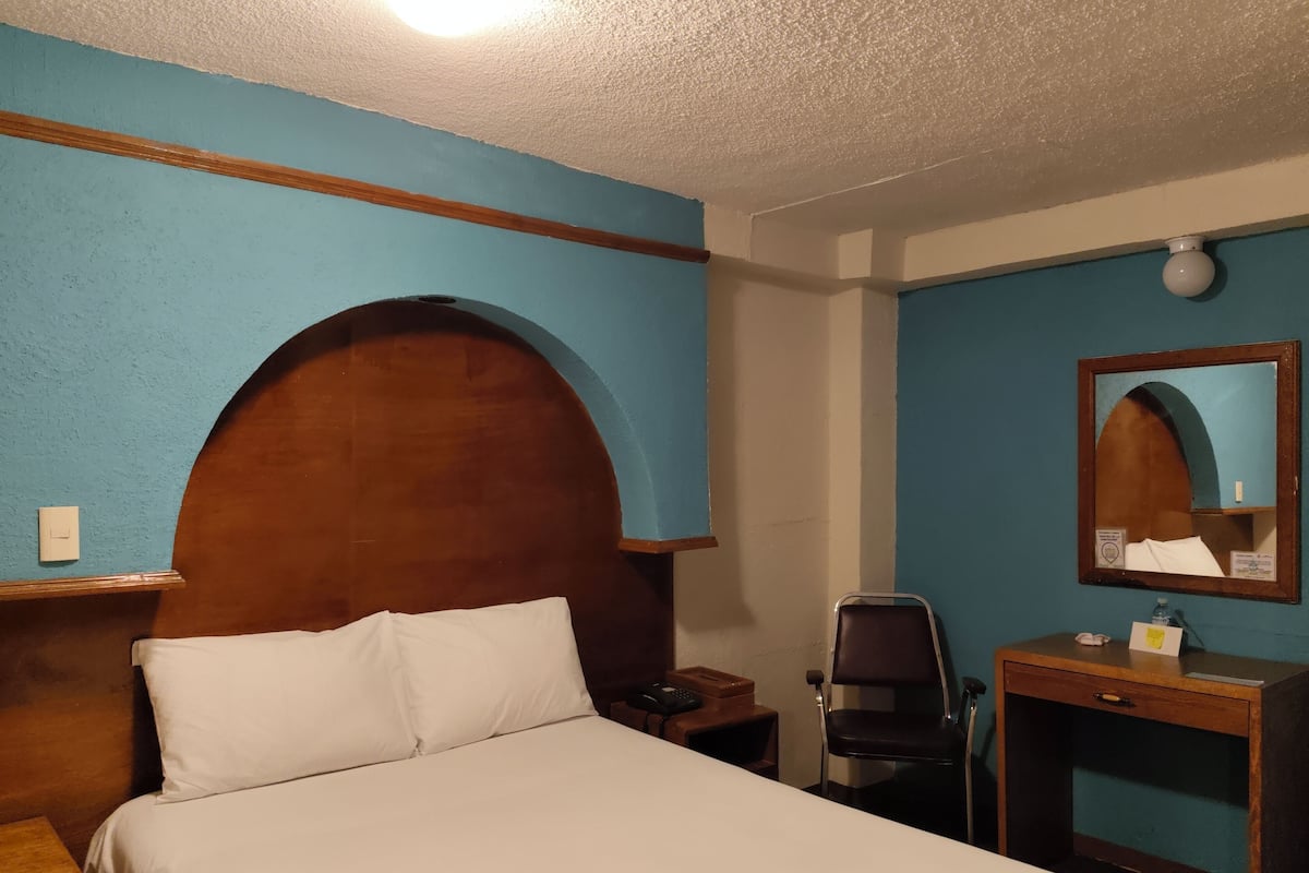 Ein Hotelzimmer mit Bett, Holz-Kopfteil, Schreibtisch mit Stuhl, Nachttisch, Spiegel und Wand mit blauer Akzentfarbe.