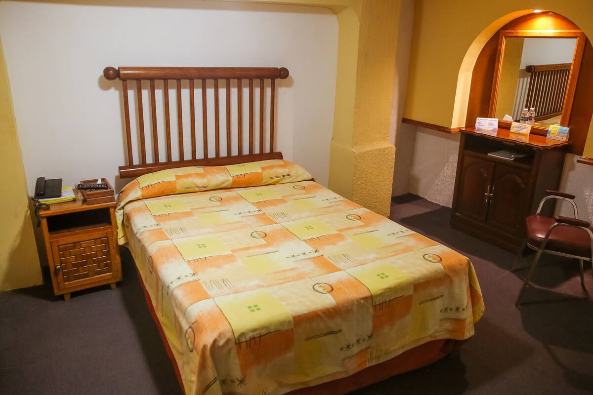 Ein Bett mit gemustertem Überwurf, ein hölzerner Kopfteil, ein Nachttisch mit Telefon und Lampe, ein Spiegel über einem Holzschrank und ein Stuhl.