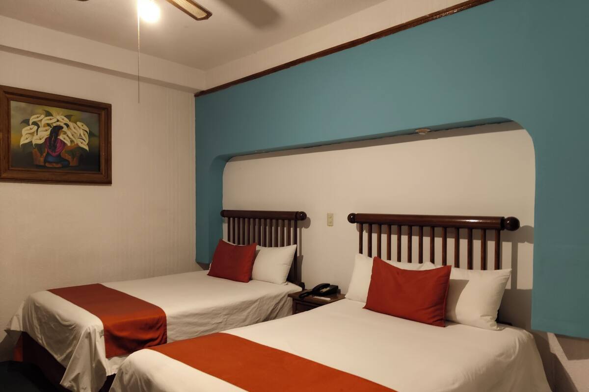 Ein Hotelzimmer mit zwei Betten, jeweils mit Holz-Kopfteilen, weißer Bettwäsche und roten Akzenten. Ein Gemälde hängt an der Wand, und ein Deckenventilator ist sichtbar.