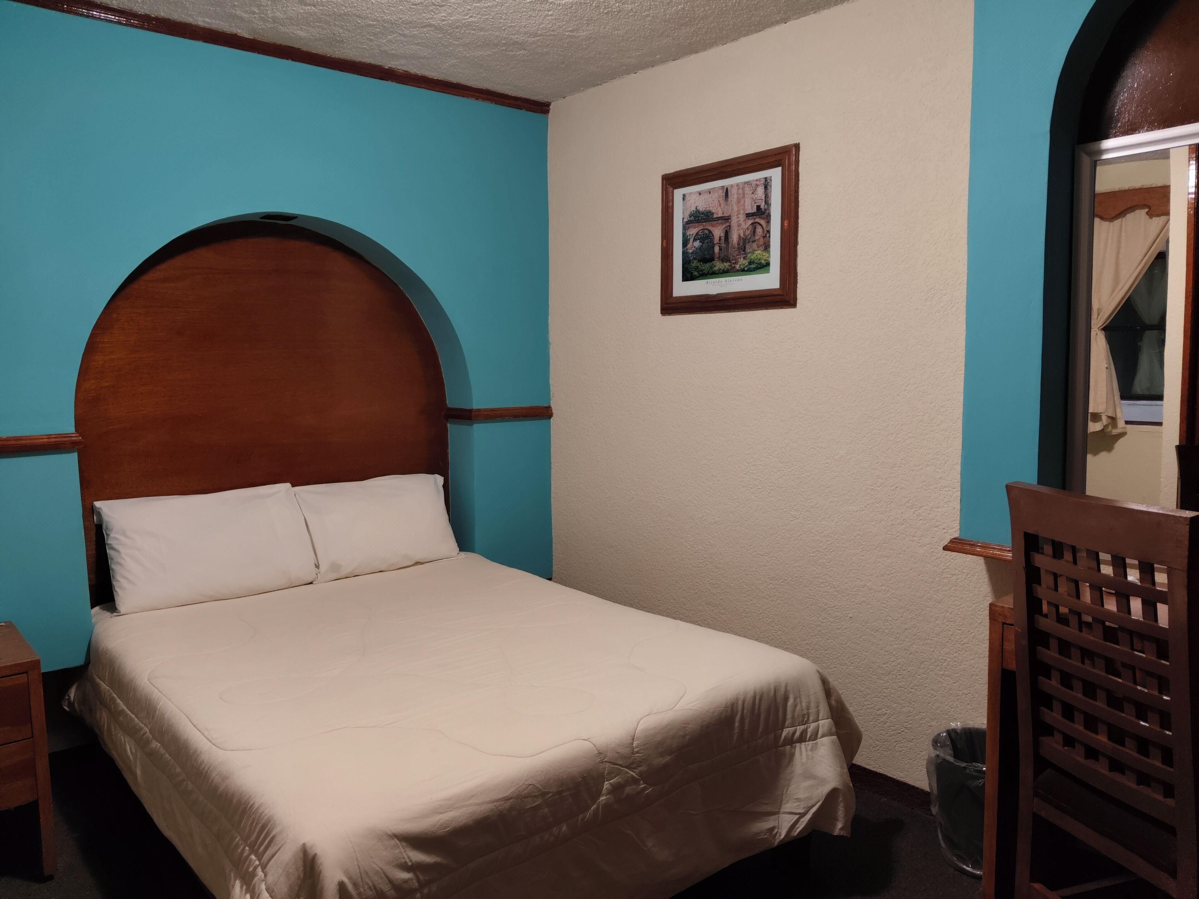 A neatly made bed in a room with a blue accent wall, a framed picture on the wall, and a wooden chair.