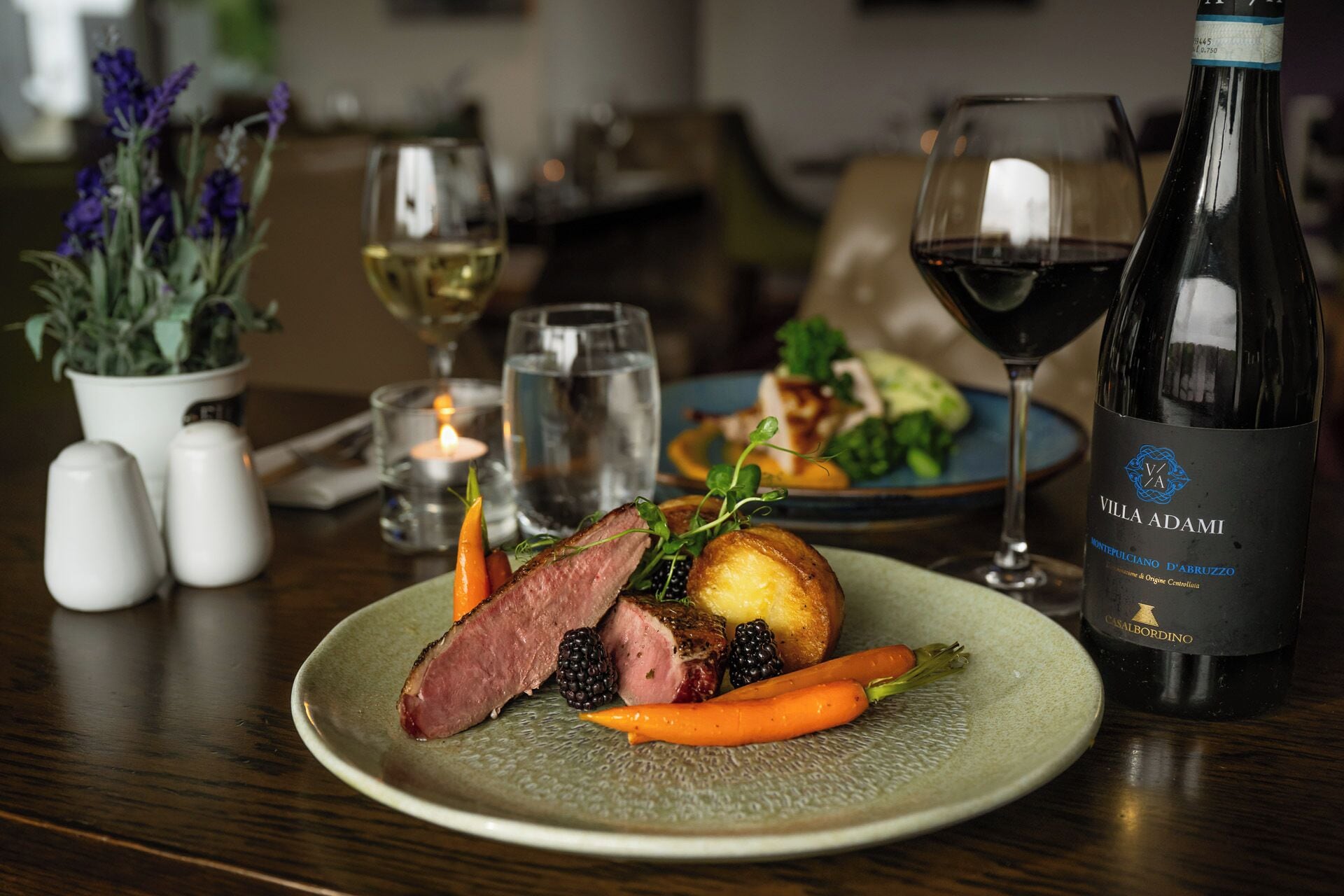 A plate of food with a bottle of wine, a glass of red wine, and a candle on a table.