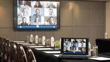 A conference room with a large screen displaying a video conference, a laptop, and several chairs arranged around a table with water bottles and papers.