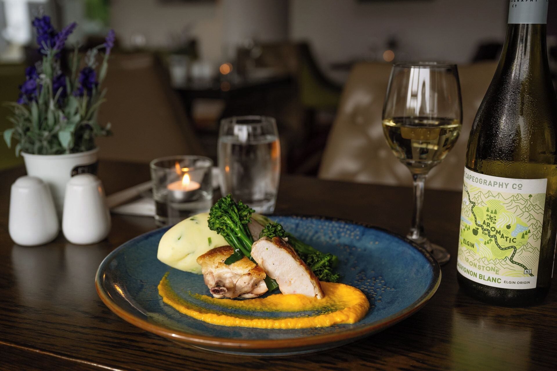 A plate with a piece of meat, vegetables, and a yellow sauce, a glass of white wine, and a bottle of wine on a table.
