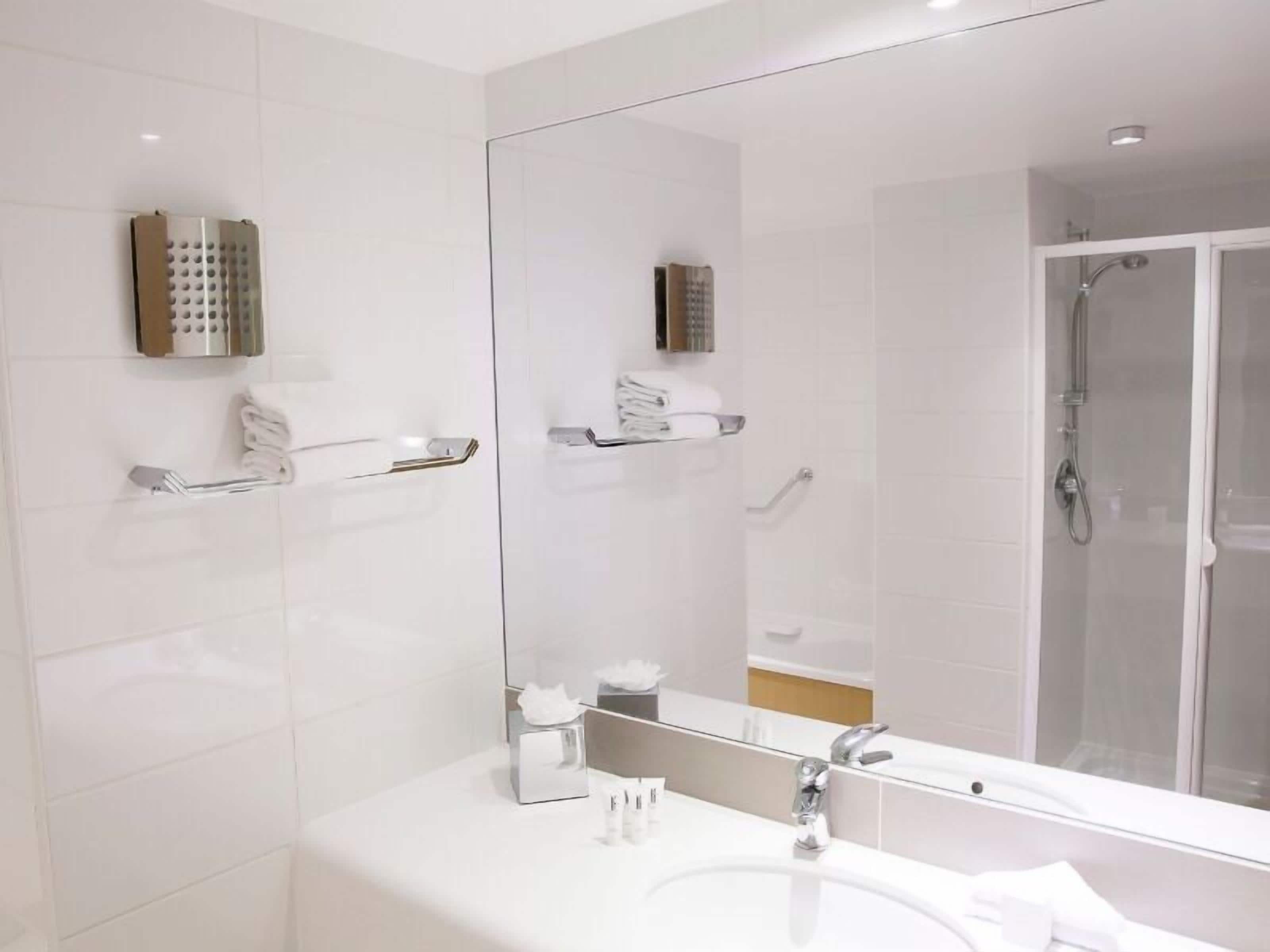 A modern bathroom with a large mirror, white tiles, a shower enclosure, and a vanity with a sink and towel rack.