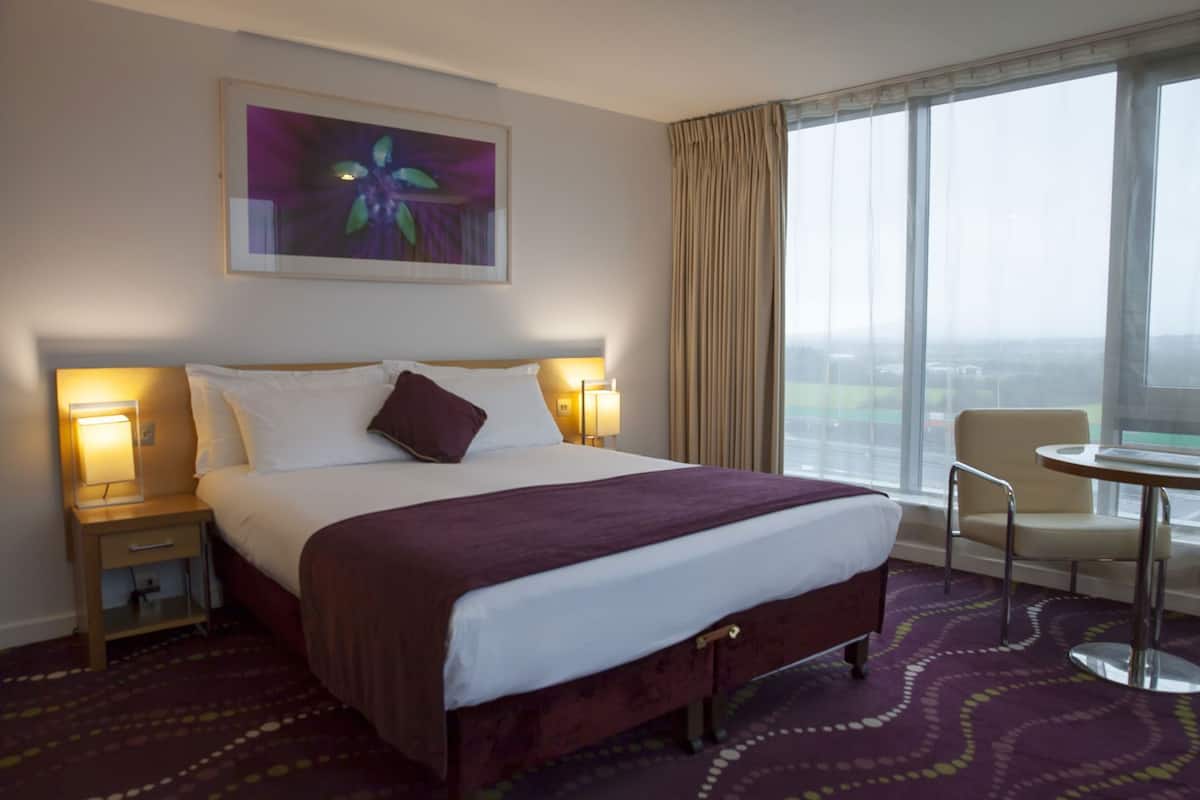 A hotel room with a large bed, two bedside lamps, a chair, a small table, and a window with curtains.