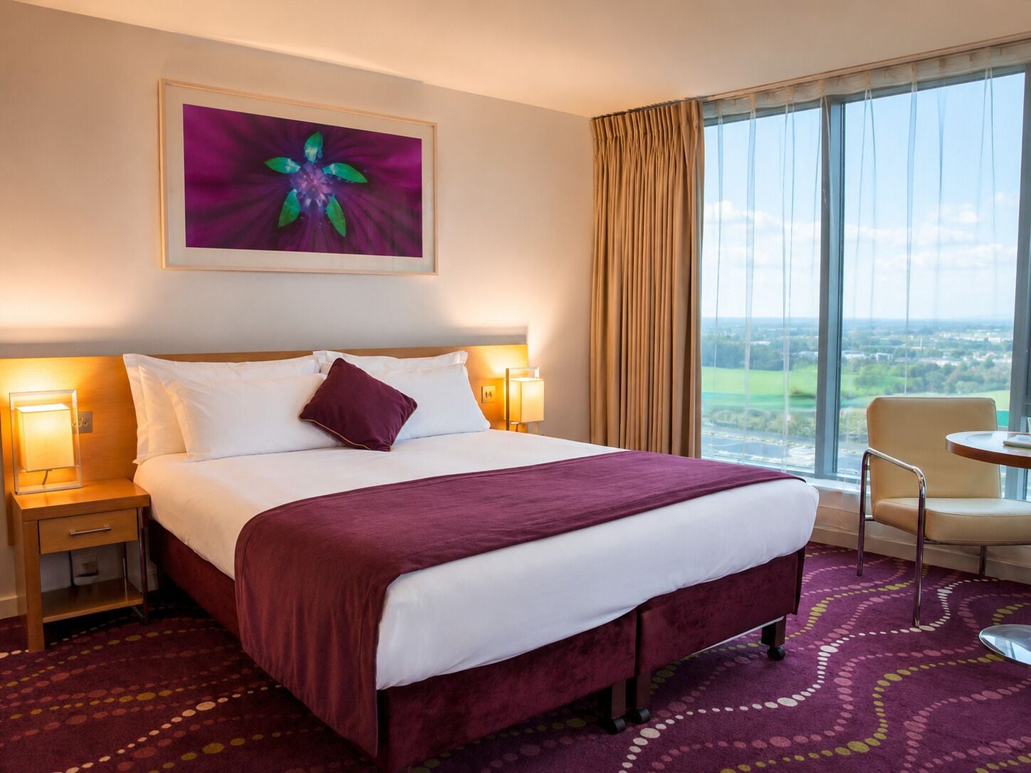 Habitación de hotel con una cama grande, vistas al campo y un área de comedor con mesa y sillas.