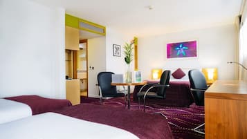 Une chambre d’hôtel avec deux lits, un bureau, deux chaises, une petite table, un vase avec une plante, un tableau encadré et une peinture abstraite colorée accrochée au mur.
