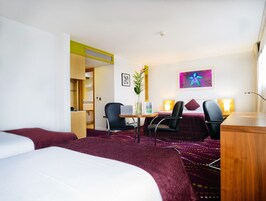 A hotel room with two beds, a desk, two chairs, a small table, a vase with a plant, a framed picture, and a colorful abstract painting on the wall.