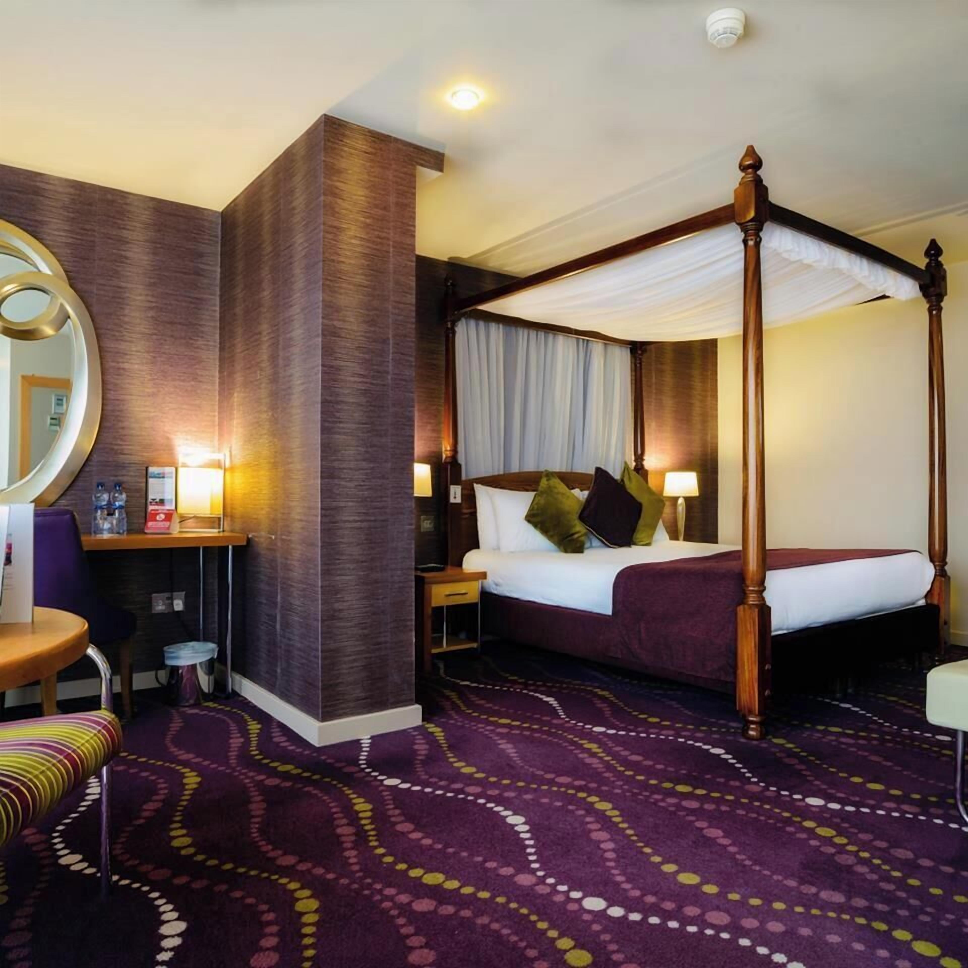 A four-poster canopy bed with a nightstand, a desk with a chair, and a round mirror.