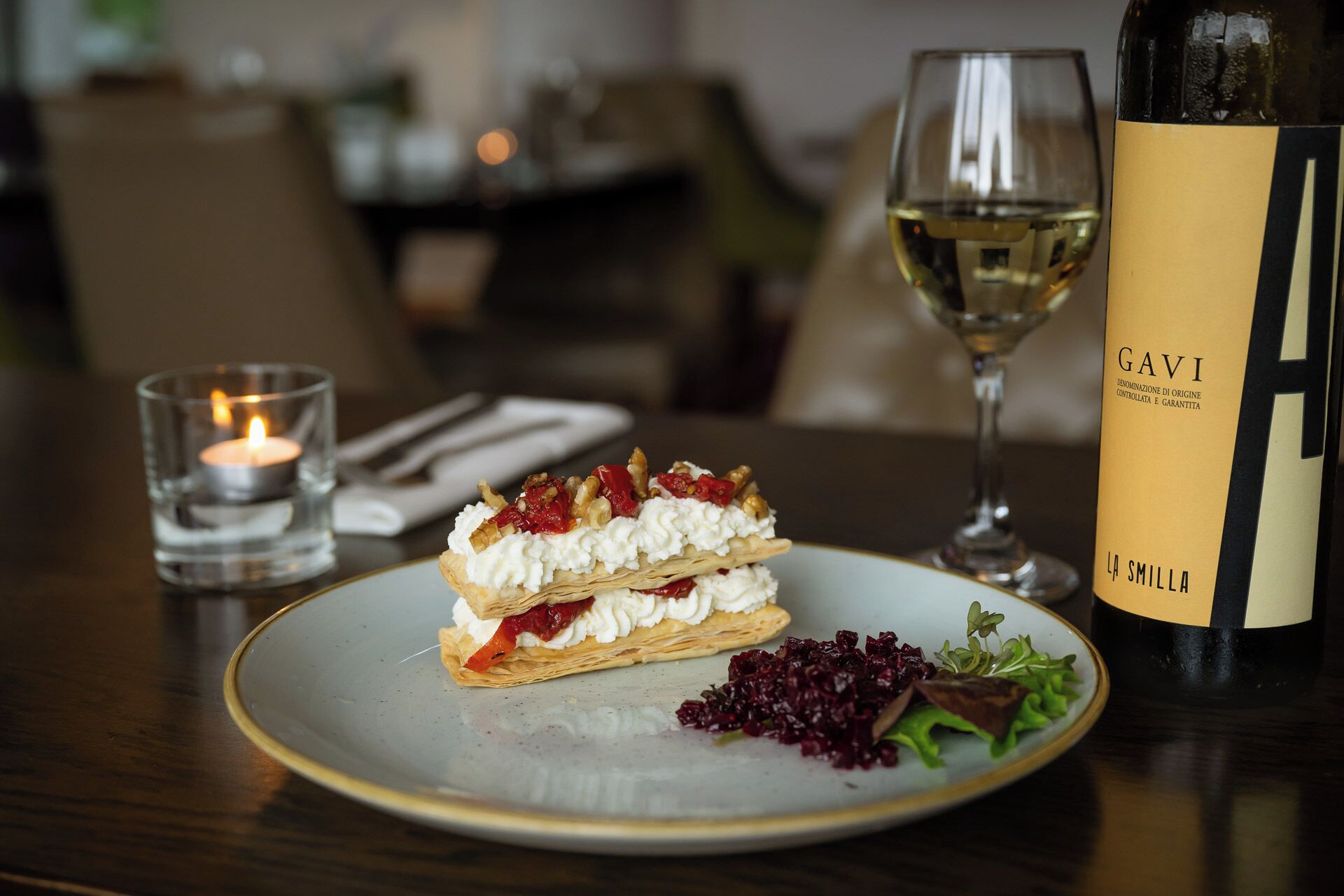 A dessert plate with a pastry, a glass of white wine, a candle, and a bottle of wine on a table.