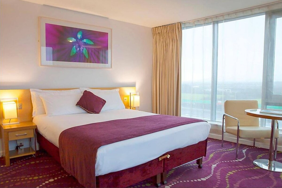 A hotel room with a large bed, two bedside lamps, a chair, a table, and a large window with curtains.