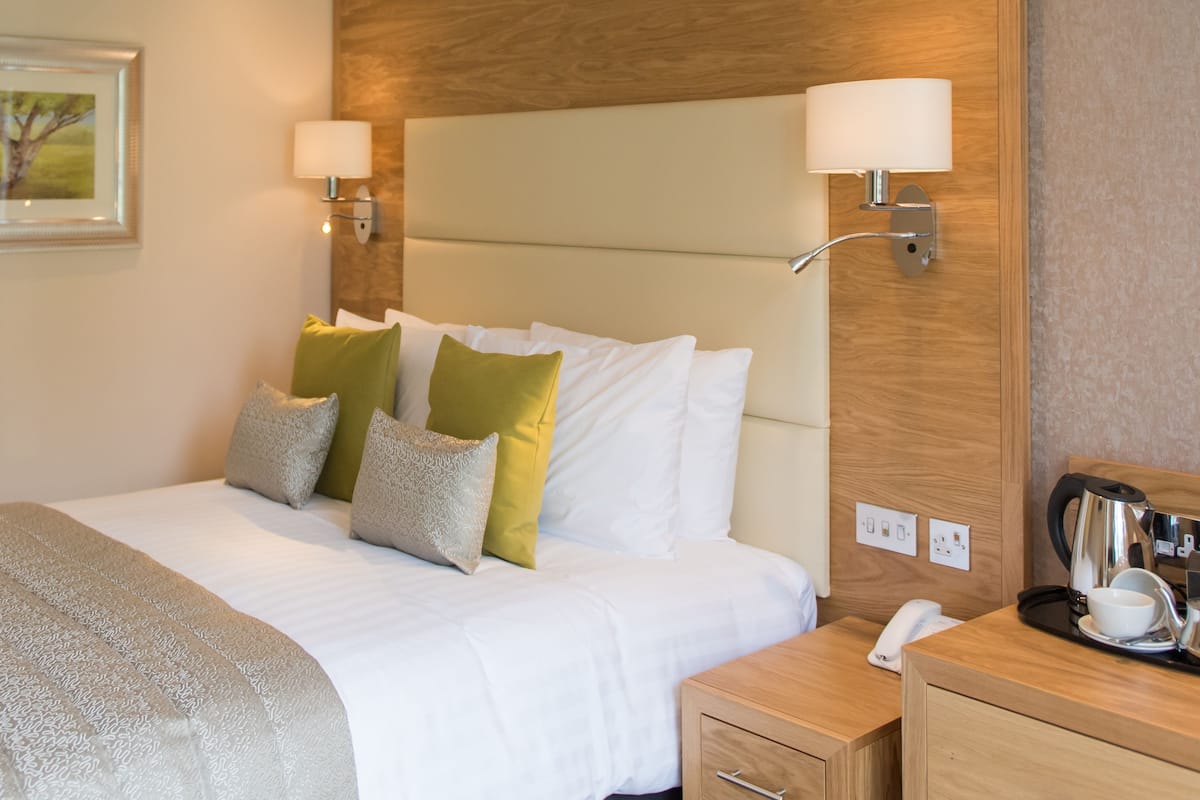A neatly made bed with white linens and decorative pillows, a wooden headboard, bedside lamps, a nightstand with a kettle, and a framed picture on the wall.