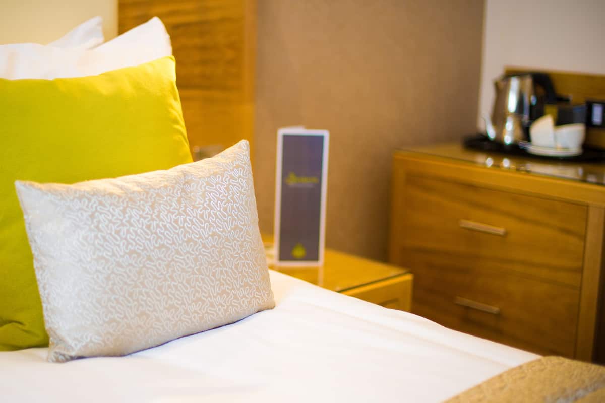 A bed with two pillows, a wooden nightstand, a coffee maker, and a box on the nightstand.