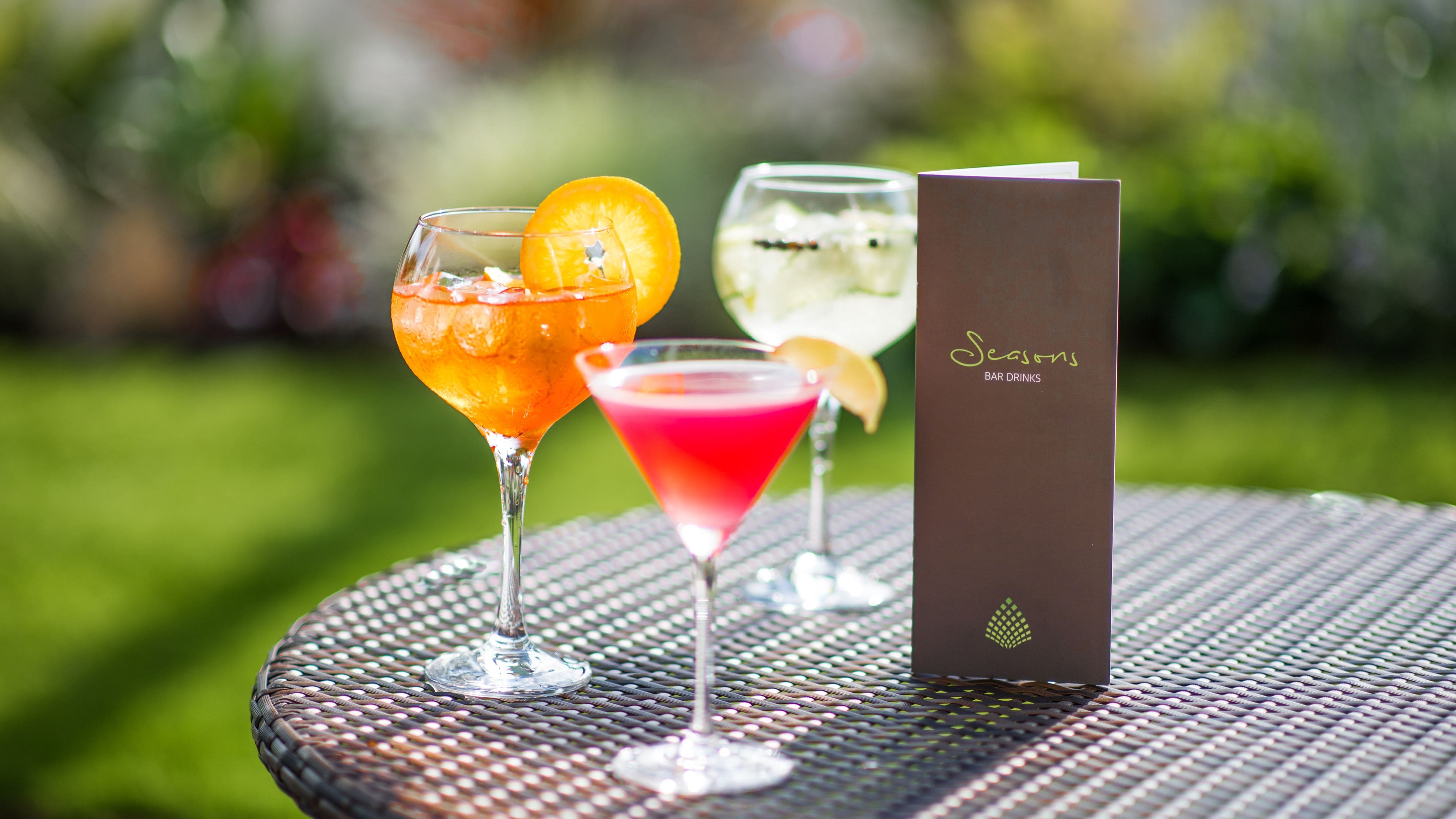 Drei Cocktails in Gläsern auf einem runden Metalltisch, neben ihnen ein Menüständer.