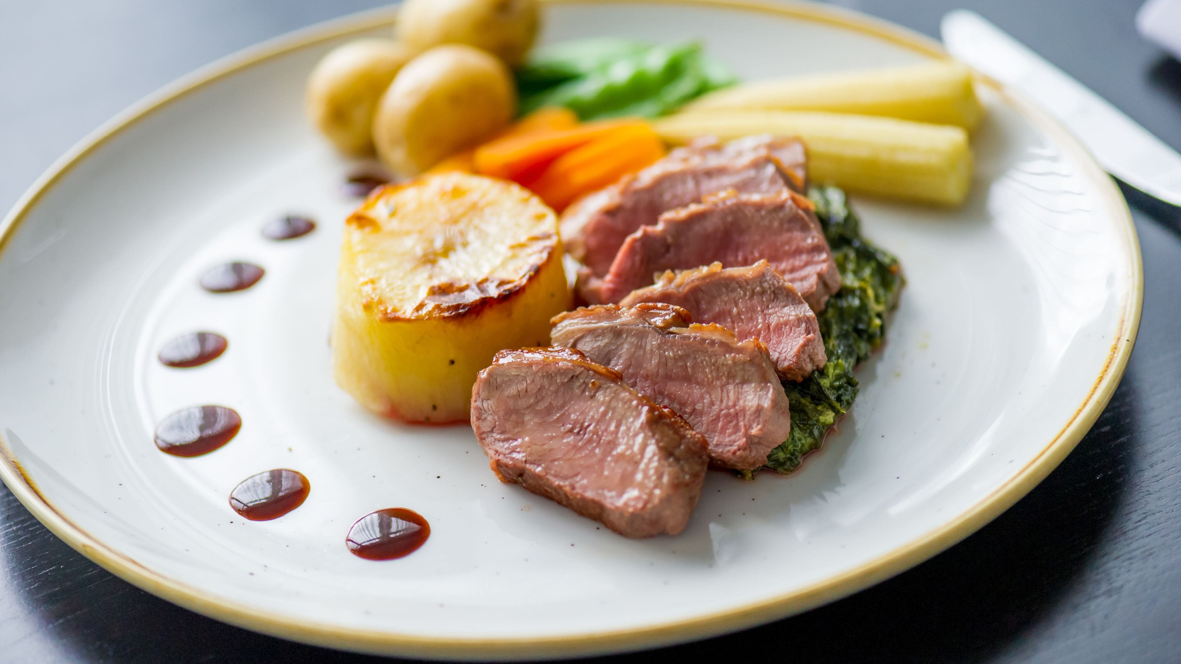 A plate with sliced meat, potatoes, carrots, and a leafy green side dish.
