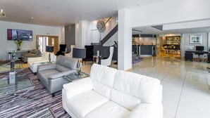 A spacious living area with a white sofa, grey and white armchairs, a glass coffee table, and a piano. There is a staircase leading to another level, and a reception desk is visible in the background. - The Park Hotel (Barnstaple)