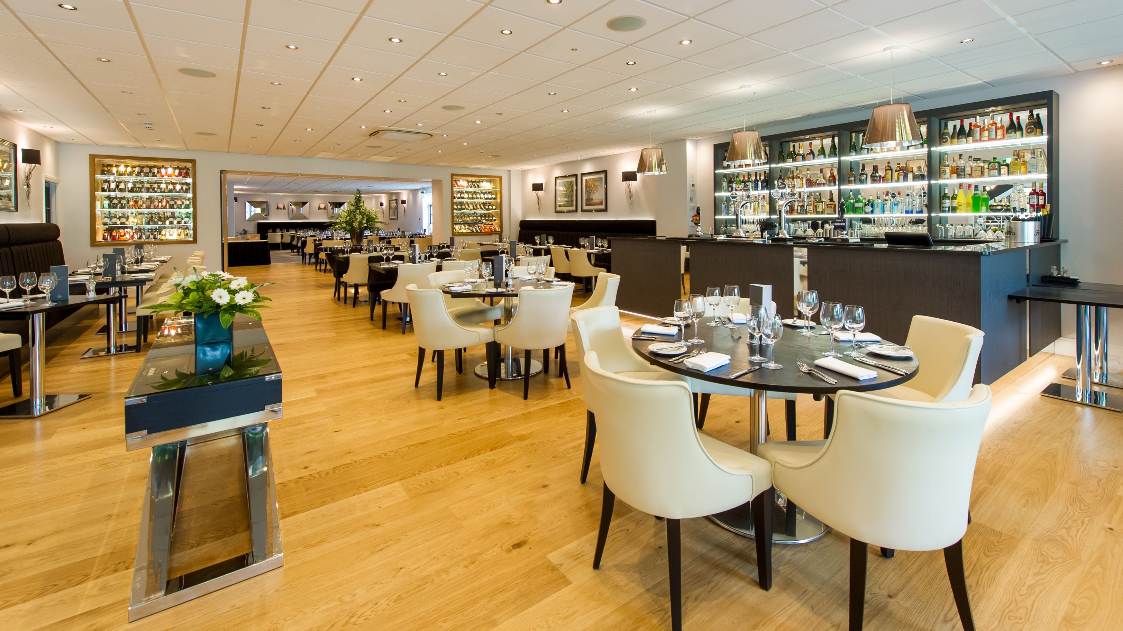 A modern restaurant with wooden floors, white chairs, and a bar area with shelves of bottles.