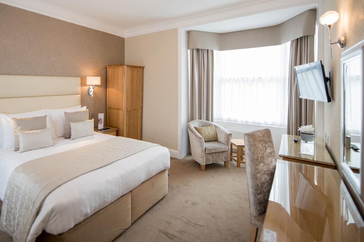 A hotel room with a large bed, a wooden wardrobe, a sitting area with a chair and a small table, a TV mounted on the wall, and a window with curtains.