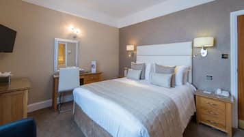A hotel room with a large bed, a desk with a chair, a mirror, and a bedside table with a lamp.