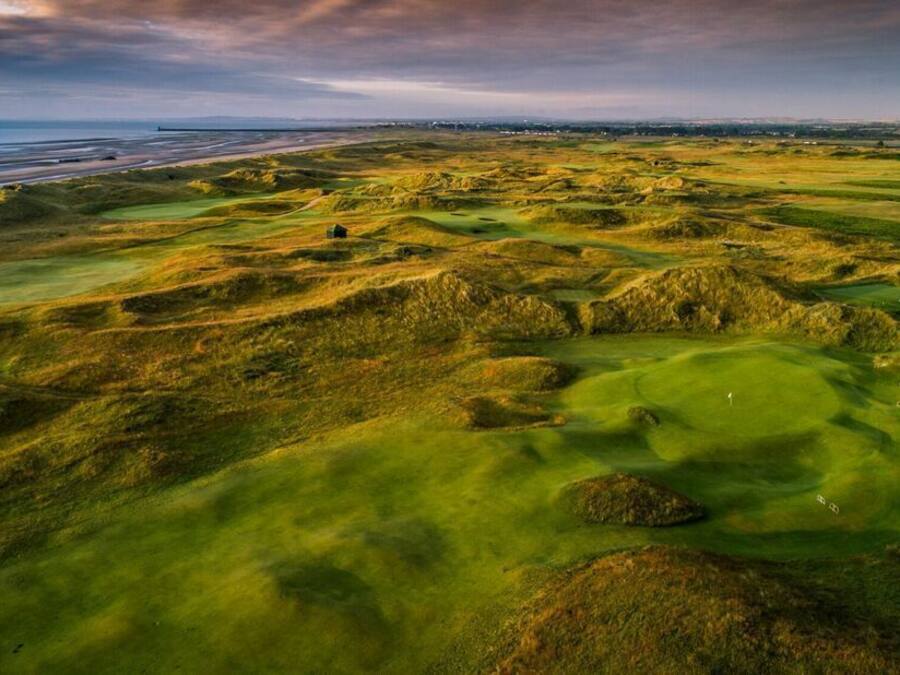 Vista aérea de un campo de golf con terreno ondulado y una costa distante.