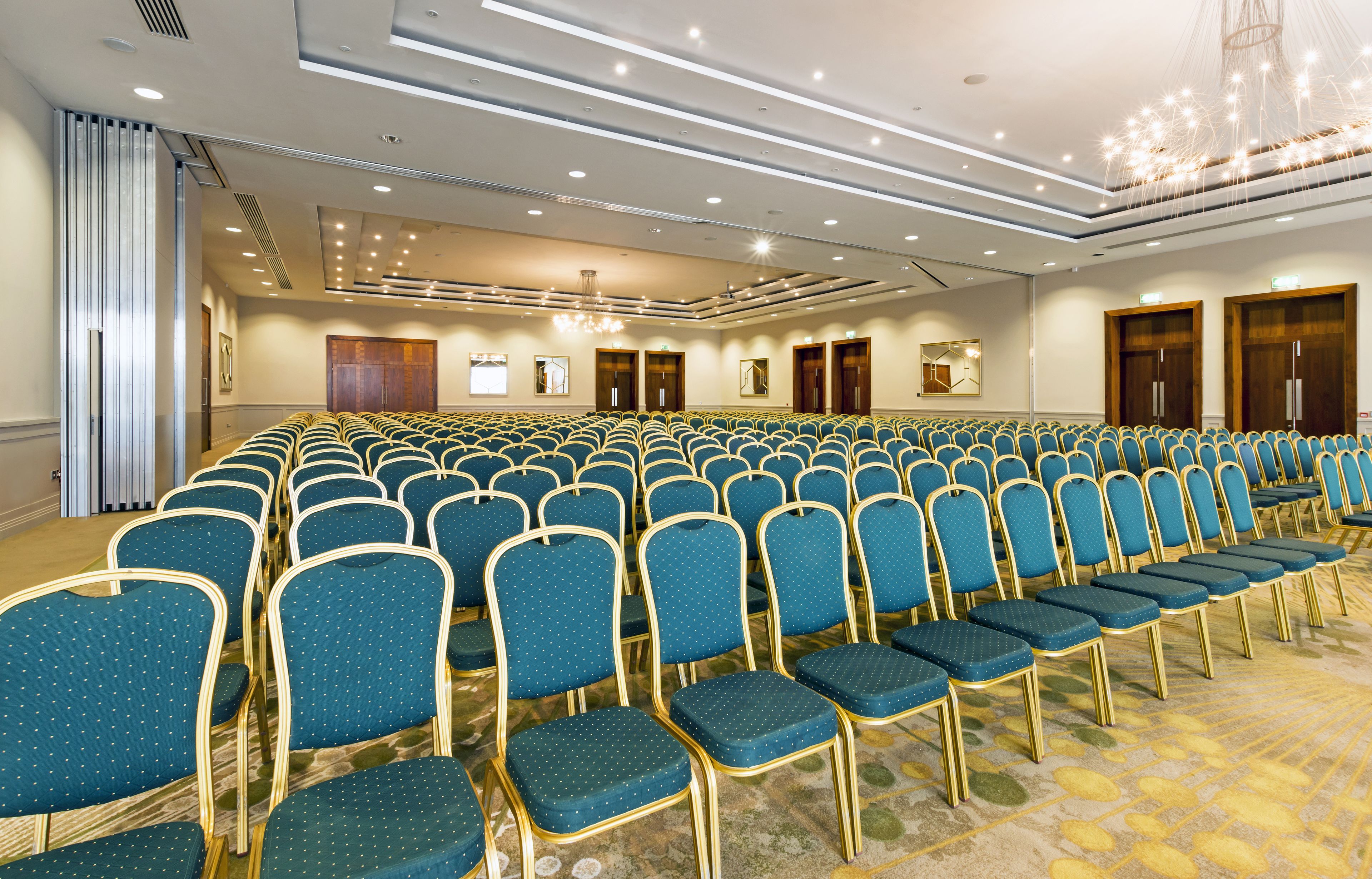 Een congreszaal met rijen blauwe stoelen, een kroonluchter en houten deuren.