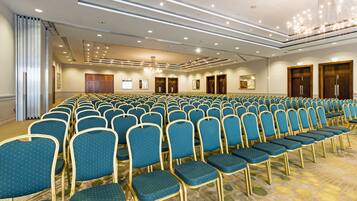 Een congreszaal met rijen blauwe stoelen, een kroonluchter en houten deuren.