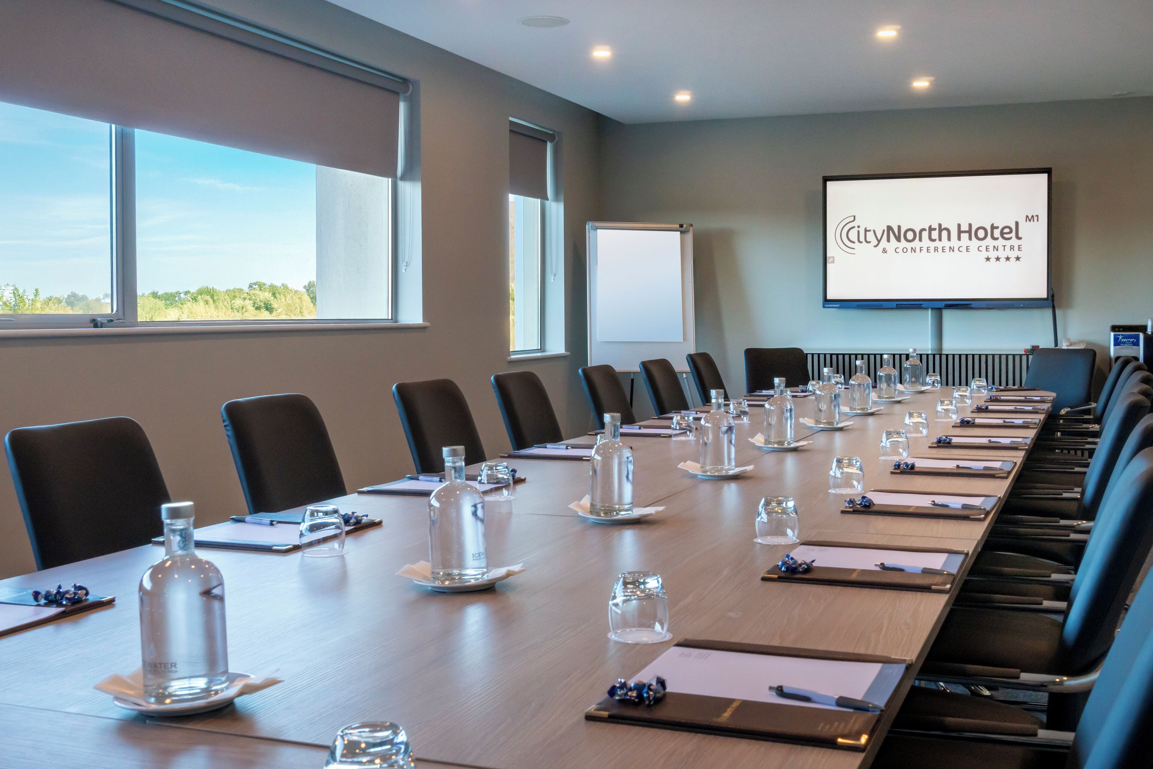 Ein Konferenzraum im CityNorth Hotel mit einem langen Tisch für Besprechungen, Stühlen, einer Projektionsleinwand und Blick ins Freie.