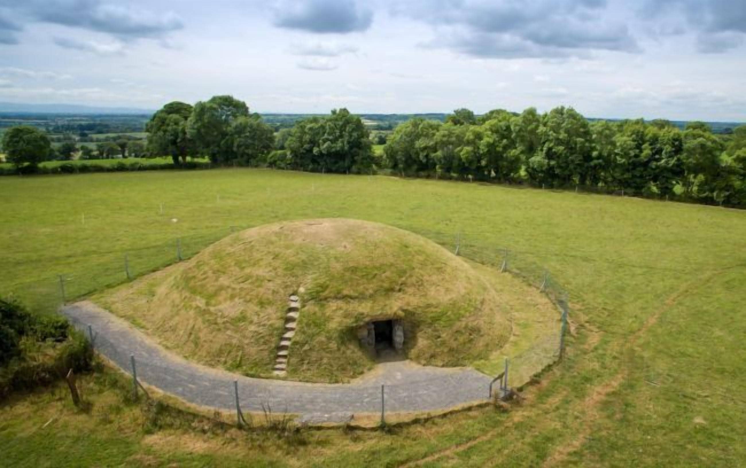 A large, grassy mound with a small entrance, surrounded by a fence and a paved path.