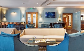 Un moderno vestíbulo de hotel con un sofá color beige, sillones azules, una mesa de centro con pasteles y un piano.