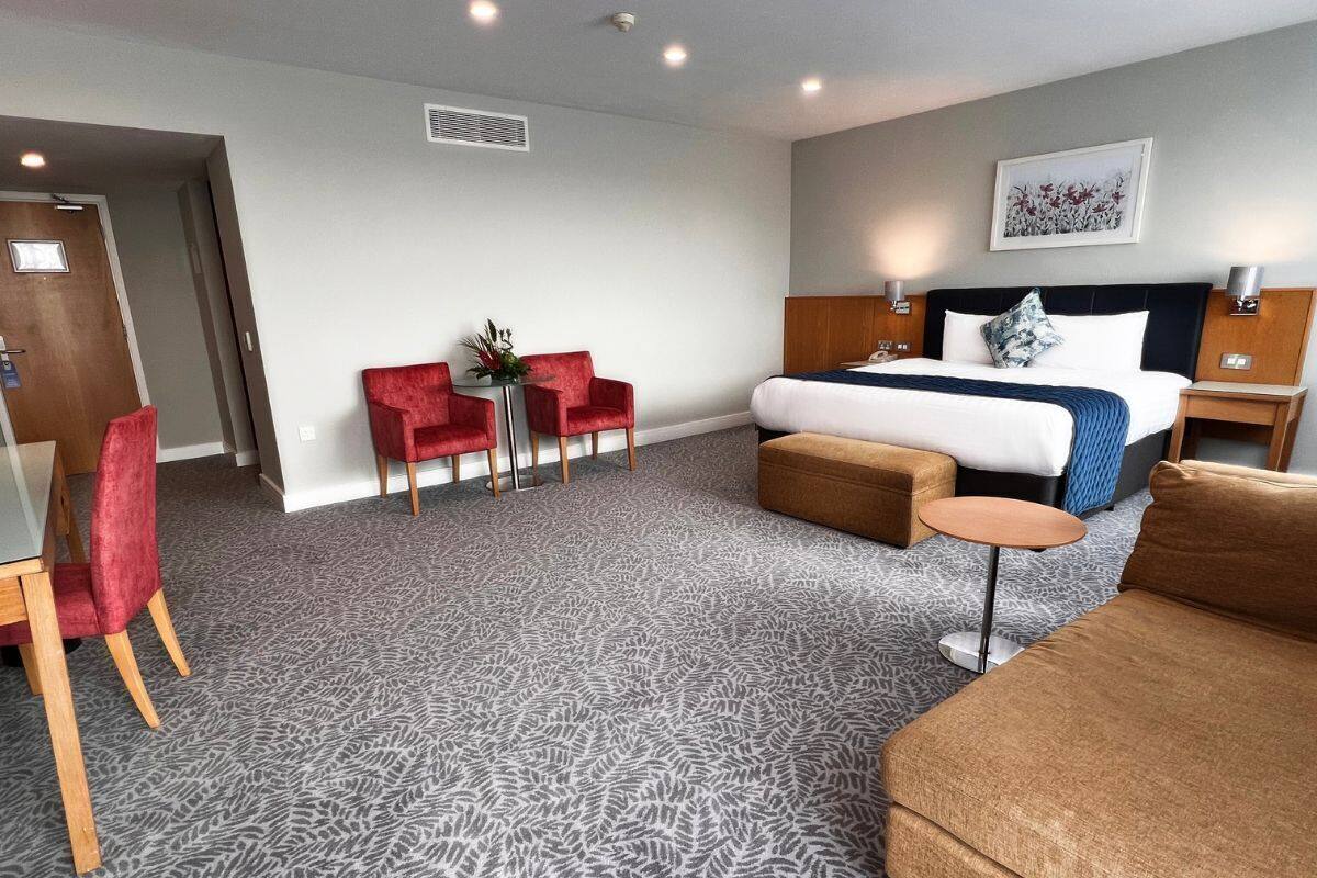 A hotel room with a large bed, a sofa, a small table, and two red chairs.