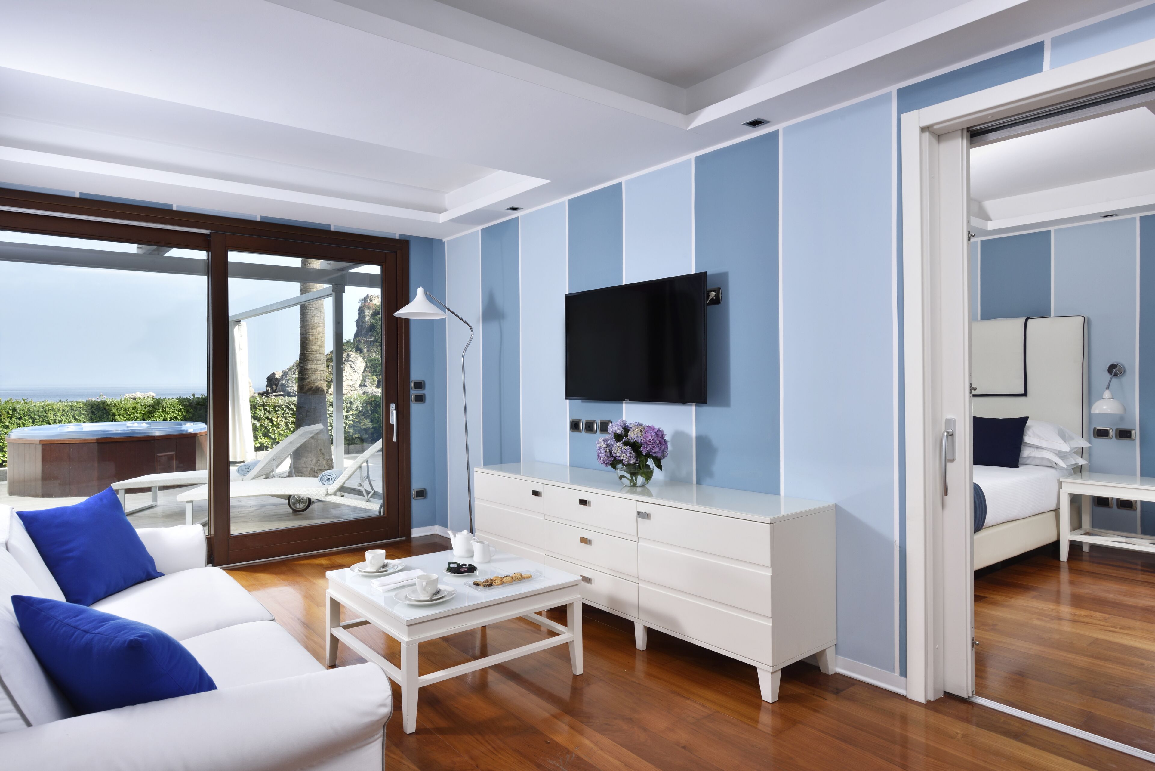 Una sala de estar moderna con un amplio ventanal corredero que da a un balcón, un sofá blanco con cojines azules, una mesa de centro blanca, un televisor de pantalla plana montado en la pared y un aparador blanco.