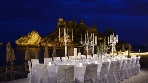 A formal outdoor dining setup with tables, chairs, and candelabras, overlooking a body of water and rocky terrain at night. - La Plage Resort (Taormina)