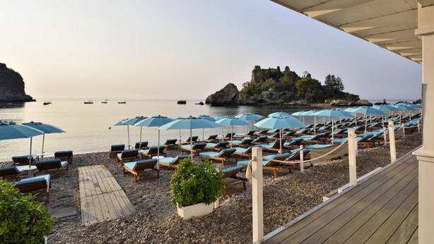 A beachside lounge area with rows of sun loungers and umbrellas, a wooden deck, and a rocky shoreline with boats in the water. - La Plage Resort (Taormina)