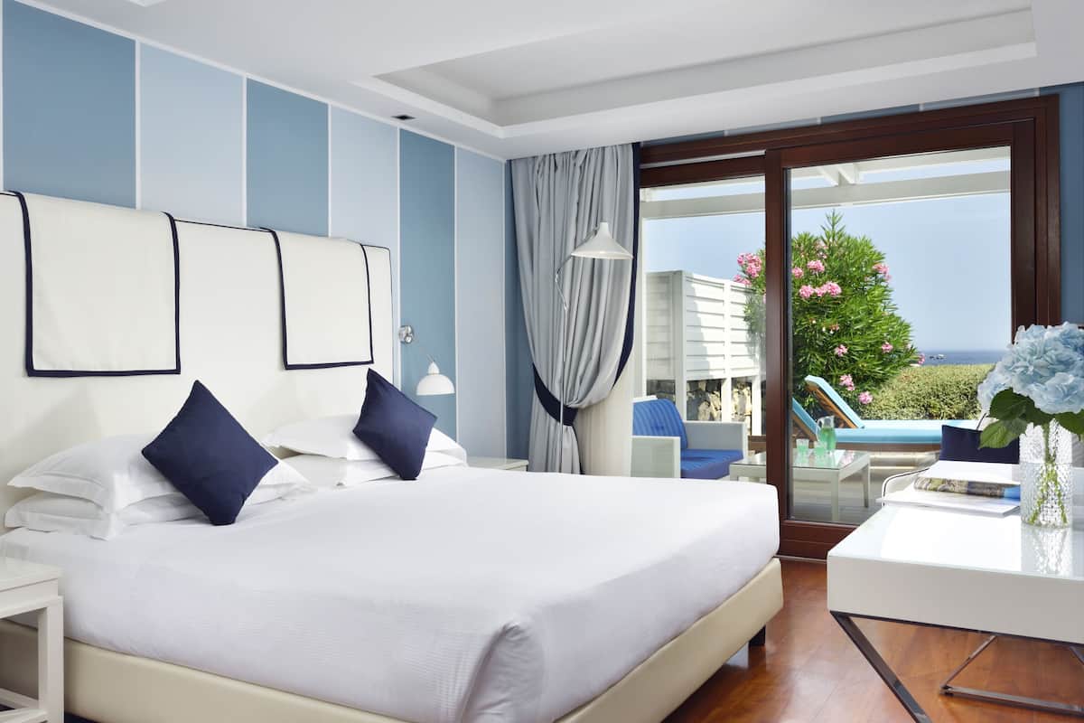A modern hotel room with a large bed, a desk, and a view of the sea.