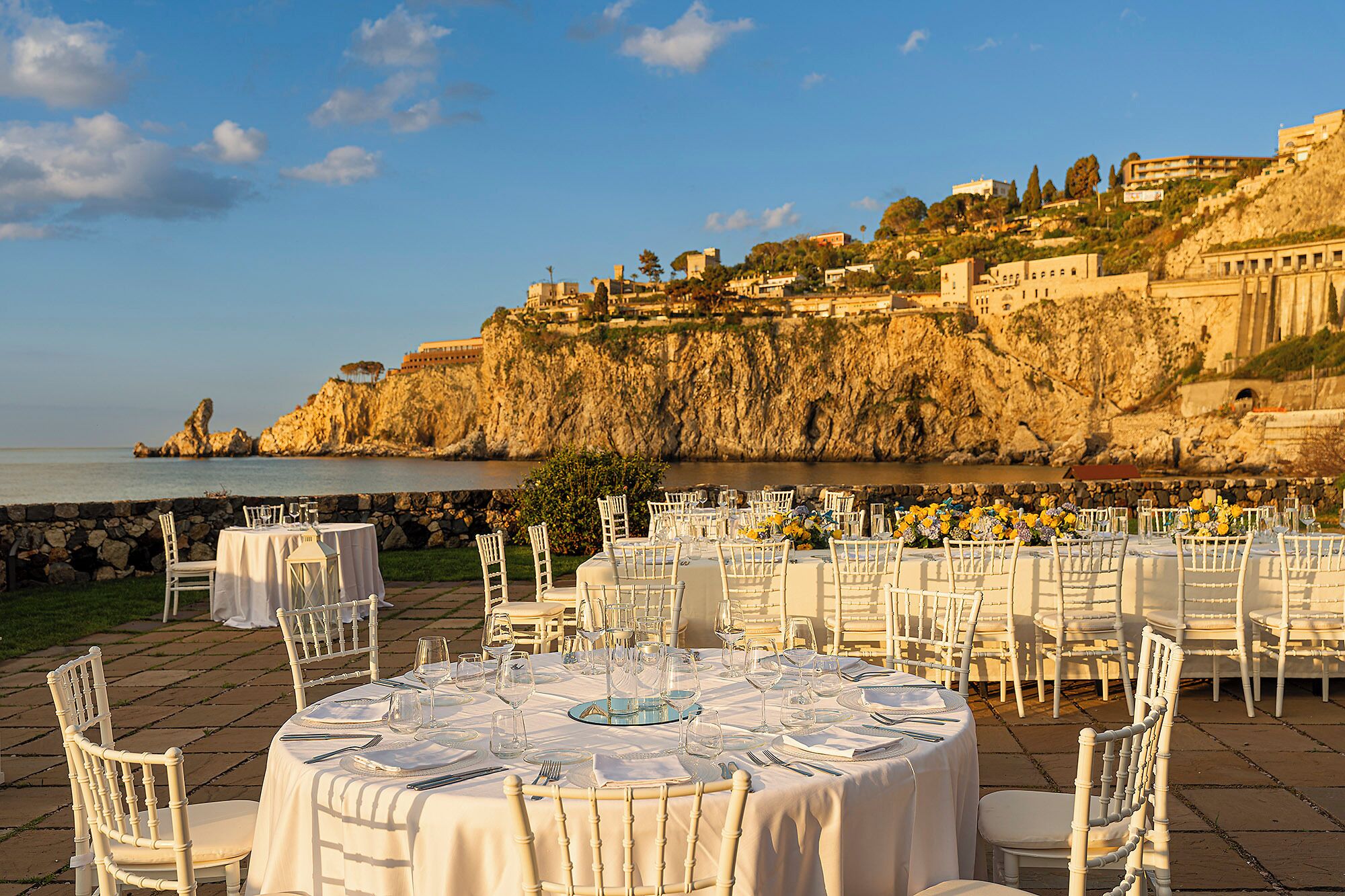 An outdoor dining area with round tables set for a meal, overlooking a coastal cliff with buildings.