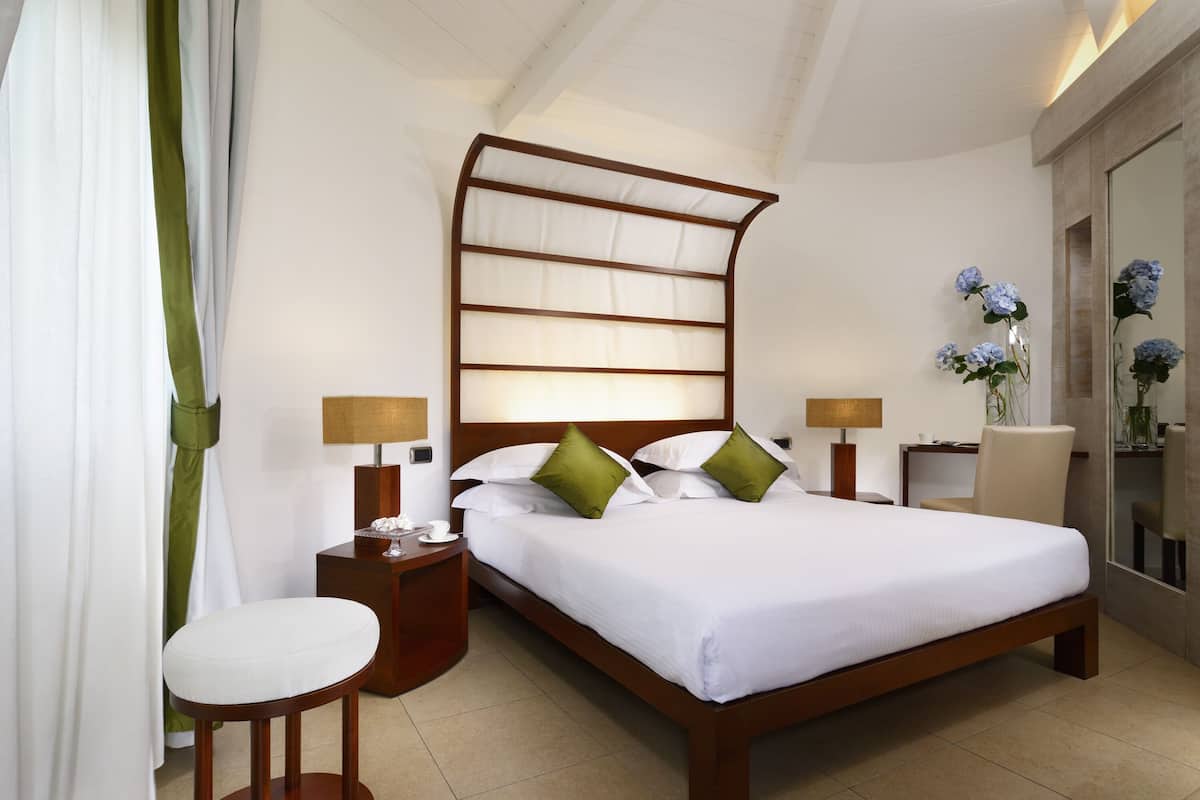 A neatly made bed with white linens and green pillows, a wooden headboard, two bedside tables with lamps, a small stool, and a mirror on the wall.