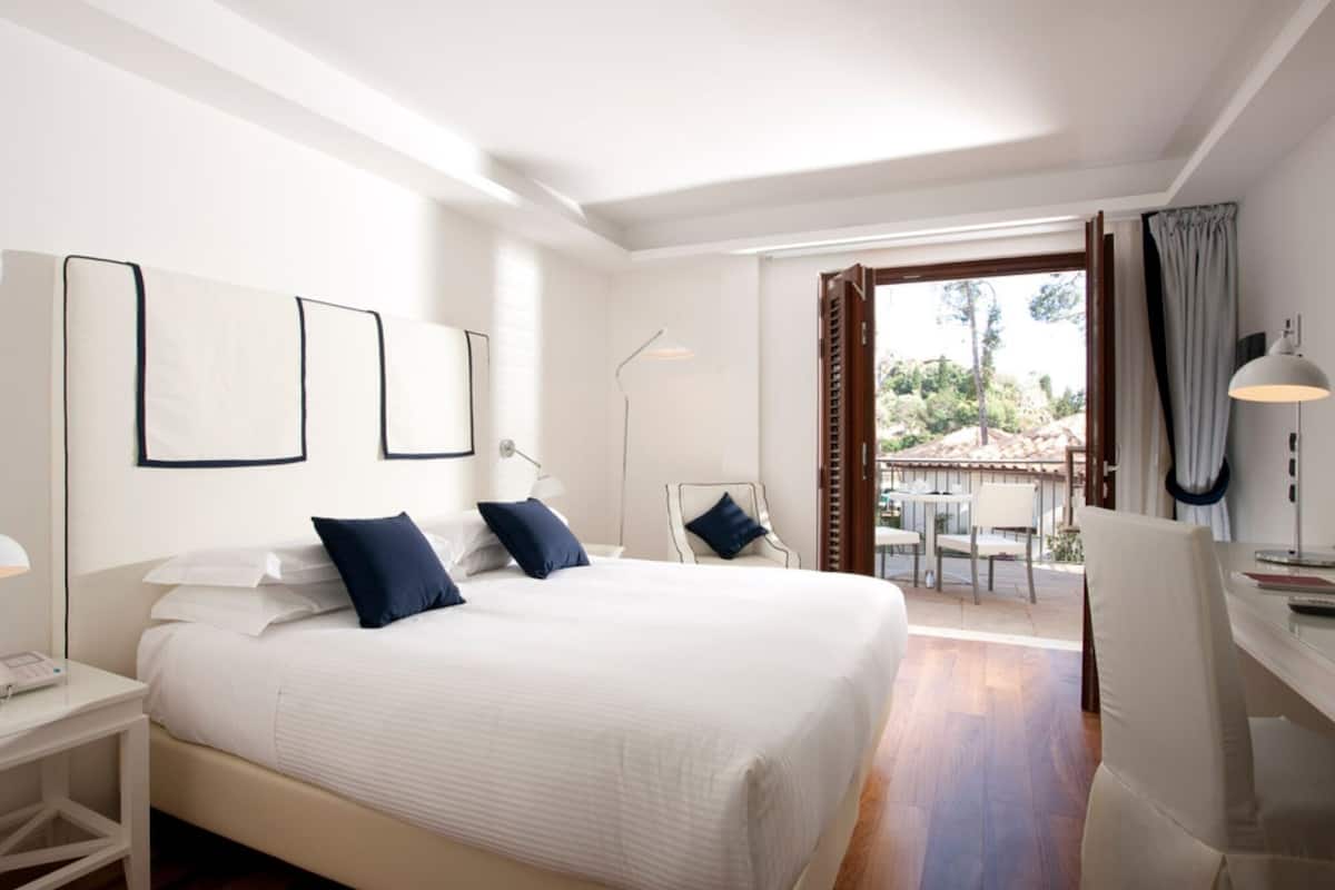 A modern hotel room with a large bed, bedside tables, a desk, and a balcony with outdoor seating.