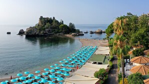 A beach with rows of blue umbrellas and lounge chairs, a rocky outcrop with ruins, and a clear view of the sea. - La Plage Resort (Taormina)