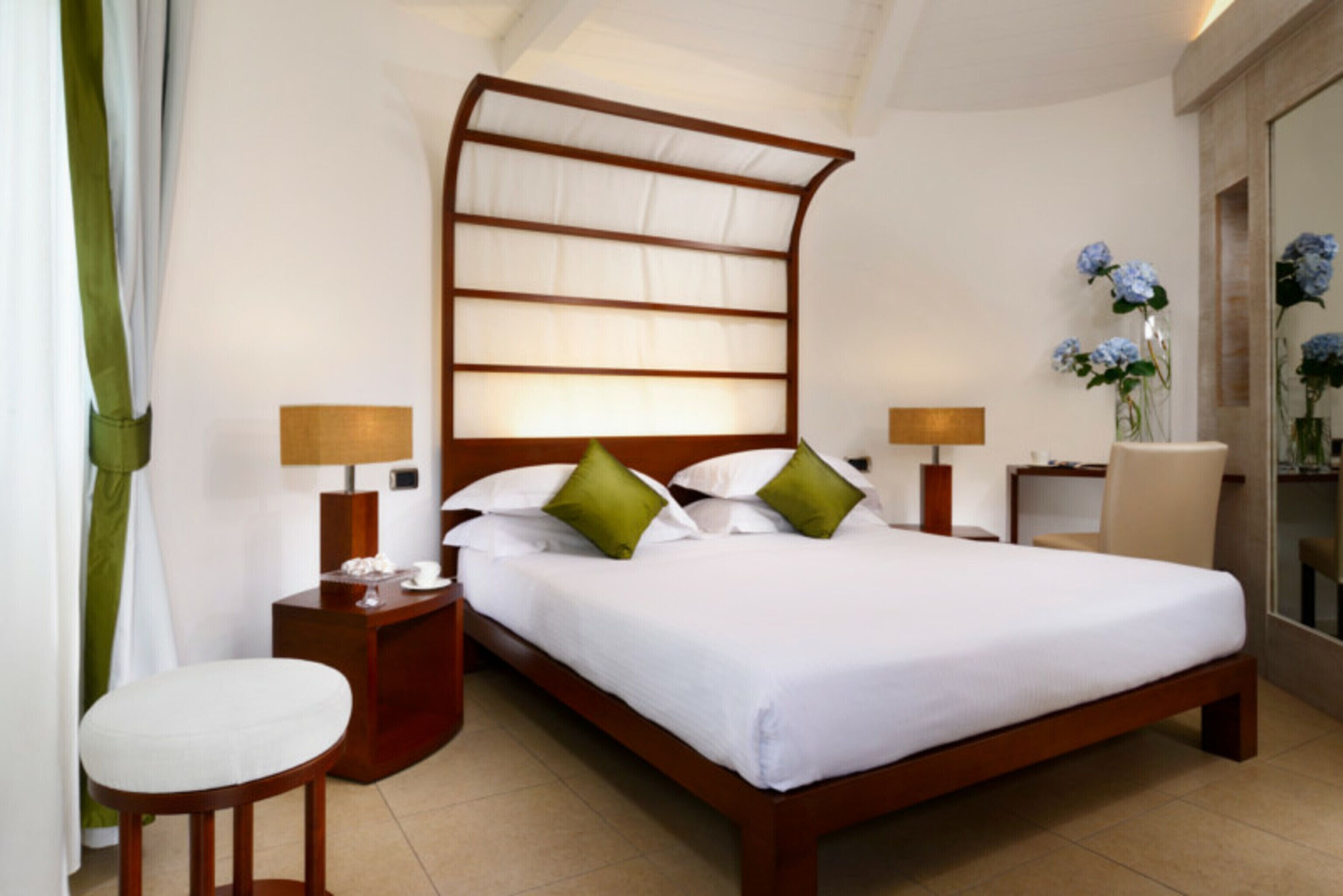 A neatly made bed with white linens and green pillows, a wooden headboard, bedside tables with lamps, and a vase with flowers on a nightstand.
